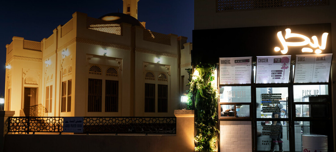 A brightly lit modern restaurant with illuminated menu boards stands beside a traditional beige mosque with intricate windows and a minaret, both seen at night under a dark sky.